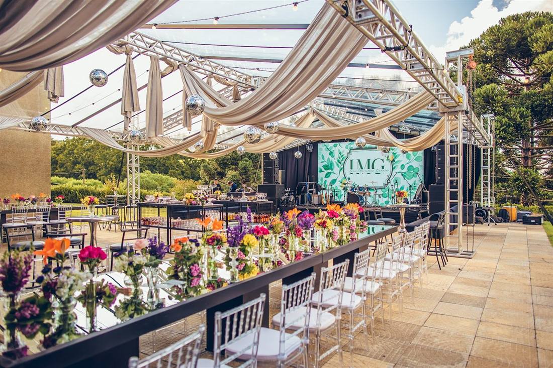 Tenda montada no gramado com mesa comprida, flores e palco, ao lado das araucárias
